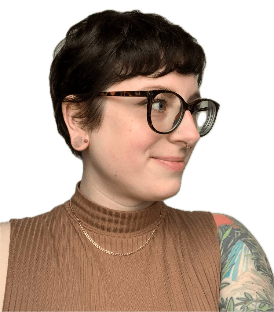 A selfie of Katie. She has short, dark hair and glasses, wearing a brown sleeveless top, with a subtle smile and a nose piercing, set against a neutral background. One arm has a large visible nature tattoo.